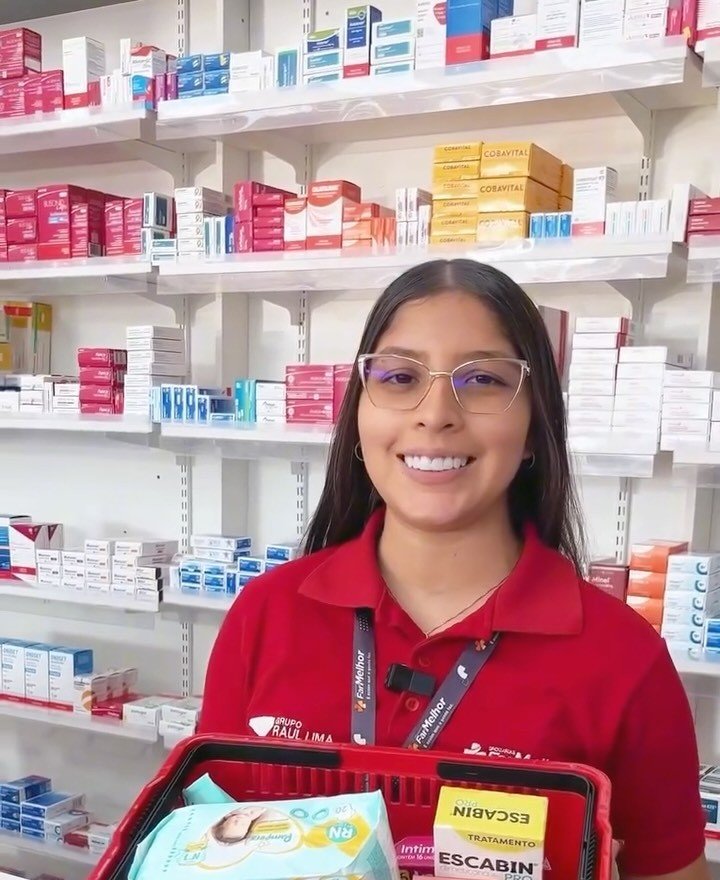 Atendente da FarMelhor, de uniforme vermelho, segurando uma cesta de compras diante das prateleiras de medicamentos da loja